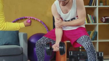 Bu video arkadaşlarınızı sıcak bir oturma odasında, birlikte spor yaparken gösteriyor. Kadın çömelirken erkek dambılları kaldırır, takım çalışmasını ve motivasyonunu teşvik eder.