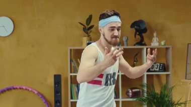 Bir adam evinin geniş oturma odasında renkli bir hula çemberiyle mutlu bir şekilde dans ediyor, neşeli ruhunu ve fiziksel uygunluğunu sergiliyor.
