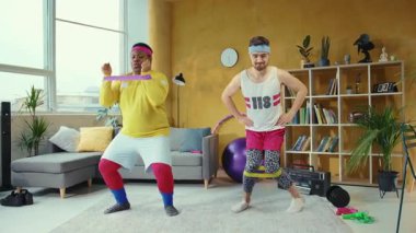 Canlı bir oturma odasında, iki adam direnç bantlarını bir fitness rutini olarak kullanır, sonra da çömelmeye geçerler. Bir erkek ve kadın birleşir, dinamiği güçlendirir.