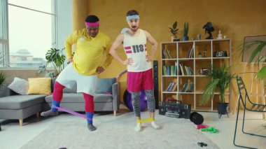 Konforlu bir oturma odasında, iki adam direniş bantlarını kullanarak dinamik bir egzersiz yapıyor, dayanıklılık, esneklik ve fitness yoldaşlığı sergiliyor.