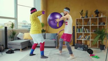 Canlı bir oturma odasında, iki adam mor pilates topuyla egzersiz yapıyor. Bir erkek ve bir kadın, takım çalışması ve eğlencesine katılıyor.