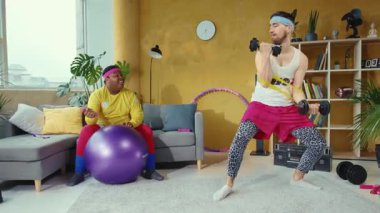 Sıcak bir oturma odasında, bir erkek dambılları kaldırırken bir kadın pilates topunun üzerinde dengede durur, çekirdek kuvveti ve aktif bir yaşam tarzı sergiler.