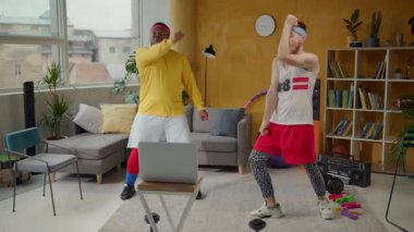 Canlı bir oturma odasında, iki adam neşeli müzik çalarken neşe ve coşkuyla dinamik dans ve fitness rutinlerine dalarlar.