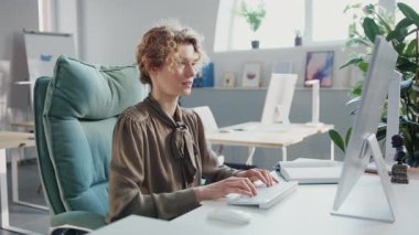 Bir kadın rahat bir şekilde bilgisayarın önünde bir masada oturuyor, görevlerine odaklanıyor ve elektronik cihazı kullanıyor.