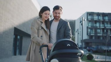 Genç bir aile göl kenarında bir bebek arabasıyla ayakta duruyor. Gün ışığında bebeği olan karı koca mutlu bir şekilde gülümsüyor. Ebeveynlik. Yavaş çekim