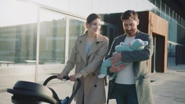 Çekici genç bir aile göl kenarında bir bebek arabasıyla yürüyor. Yeni doğmuş bebeği tutan adam. Güneş ışığında gülümsüyor. Ebeveynlik. Yavaş çekim