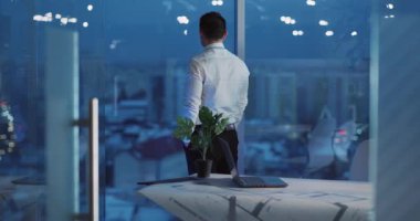 Düşünceli bir profesyonel gece şık ve modern bir ofisin penceresinden dışarı bakar, fikirleri derinden yansıtır.