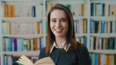 Genç bir kadın, kitaplarla dolu uzun raflarla çevrili, rahat bir kütüphanede kitap okumaya dalmış.