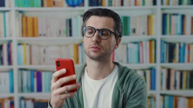 Genç bir adam, kitaplıklarla dolu canlı ve renkli bir modern kütüphanede akıllı telefonuna odaklanmış.