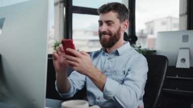 Modern, çağdaş ofis ortamında bir masada özenle çalışan bir adam akıllı telefonunu kullanıyor.