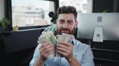 Çok neşeli bir adam, iyimserlikle dolu, gösterişli ve modern bir ofis ortamında gururla para yığını sunar.