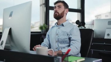 Sakallı bir adam, şık ve modern ofis ortamında bir masaüstü bilgisayarı üzerinde çalışırken derinden odaklanır.