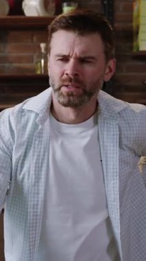 Sıradan kıyafetler giymiş bir adam, rahat bir iç mekanda otururken düşünceli bir yüz ifadesi sergiliyor.