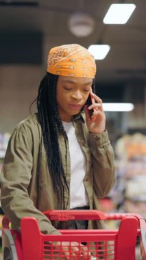 Cep telefonuyla konuşan Afro-Amerikalı bir kadın alışveriş arabasının içine yiyecek koymakla meşgul. Müşteriler. Tüketim. Rutin kavram.