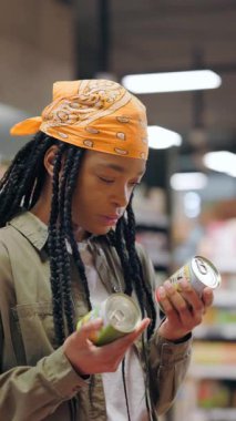 Afro-amerikan genç, şık, sağlıklı görünümlü bir kadın, yiyecek satın alıyor, konserve ürünleri inceliyor, iki öğe arasında seçim yapıyor. Akıllı alışveriş. Tüketim. Market..