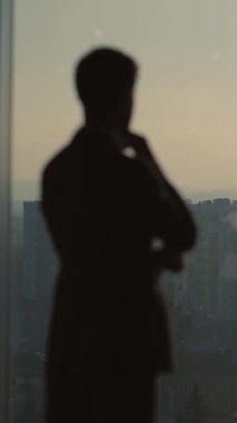İyi giyimli bir adam büyük bir pencerenin yanında duruyor ve cep telefonuyla konuşuyor. Silueti derin düşüncelere daldığını ve profesyonellik yaydığını gösteriyor.