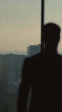 Bir video, takım elbiseli bir adamı pencerenin yanında, düşünceli bir şekilde şehri izlerken gösteriyor. Işık, hırsı ve şehir yaşamını yansıtan bir siluet yaratır.