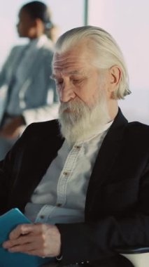 Samimi bir ortamda, sakallı yaşlı bir adam okumaktan ve tablet kullanmaktan hoşlanır. Doğal ışık ve mavi bir klasörle rahatça oturuyor. Düşüncelerini yazıp düzenlediğini söylüyor.