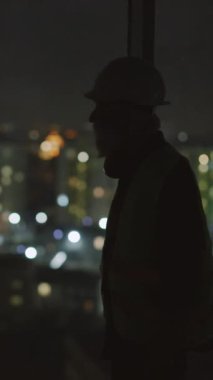 Loş bir ortamda, sert şapkalı bir adam düşünceli bir şekilde pencereden dışarı bakar ve gölgelerin arasında yalnız varlığını vurgular.