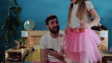 Kızıyla oynarken akıllı telefon kullanan sıkılmış genç bir babanın portresi. Neşeli şirin kız babasının etrafında dans eden peri gibi üzgün üzgün üzgün yatakta oturmuş eğleniyormuş gibi yapıyor..