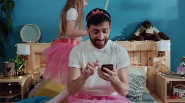 Akıllı telefon kullanarak prenses gibi giyinmiş sosyal, güler yüzlü, genç bir baba. Kızı sihirli bir değnekle yatakta oynayan bir peri oynuyor..