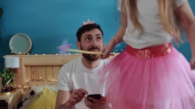 Genç, yorgun, tacını takmış akıllı telefondan kızına gülümseyen sihirli değnekle yatakta dans eden süslü elbiseli genç bir baba. Çocuklarla evde parti.