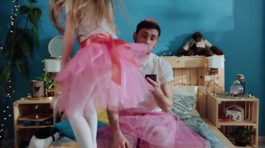 Yatakta oturup yüzüne dokunarak akıllı telefon kullanan yorgun ebeveyn portresi. Yakışıklı genç adam çocuklarla oynamaktan yoruldu. Güzel kız dans ediyor ve yatakta eğleniyor.