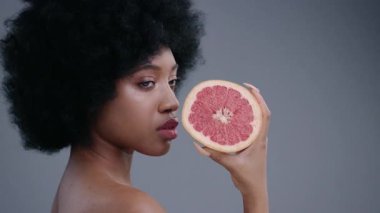 Zarif Afro-Amerikalı genç bayan elinde sulu bir greyfurtla kameraya bakıyor. Gri arka planda turunçgil meyvesi tutan neşeli güzel bir kızın profili..