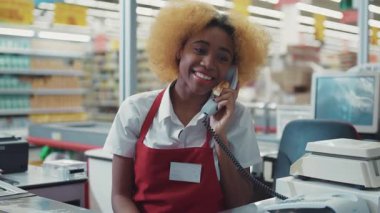 Gülümseyen Afrikalı Amerikalı kadın süpermarkette çalışıyor. Para masasında çalışıyor. Telefonla konuşuyor.