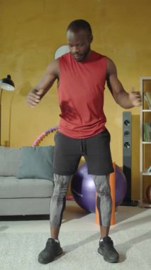 Formda bir adam evde çeşitli egzersizler sergiliyor şık spor kıyafetleri ve çeşitli spor aletleri sergiliyor.