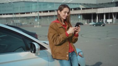 Genç bir kadın, şehir ortamında park halindeki bir arabanın yanında akıllı telefonuyla mesajlaşmaktan hoşlanıyor..