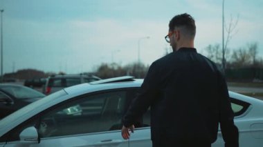 Bir adam bulutlu bir gökyüzünün altında, geniş bir açık hava otoparkında, gösterişli beyaz bir arabaya doğru yürüyor.