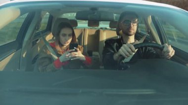 Genç bir çift, araba kullanırken bir kişinin akıllı telefonuyla meşgul olduğu bir arabada görülebilir.