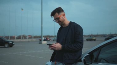 Bir adam geniş ve boş bir otoparkta arabasına yaslanırken aktif olarak mesajlaşıyor.