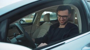 Bir adam arabasının sürücü koltuğunda oturur ve araç hareket halindeyken tamamen akıllı telefonuna odaklanır.