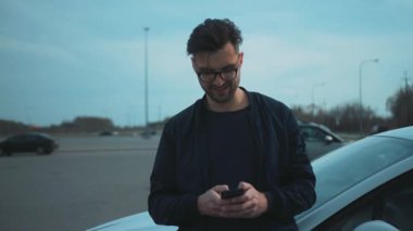 Meşgul bir otoparkta arabasına yaslanmış şık bir genç adam akıllı telefonunu rastgele kontrol ediyor.