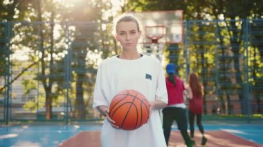 Çekici bir sahnede, dışarıda arkadaşlarıyla çevrili canlı bir kız elinde basketbol topu tutuyor.