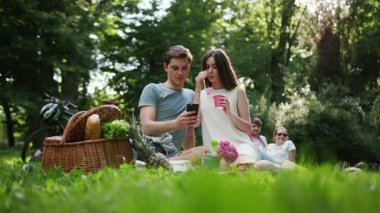 Gülümseyen mutlu genç çift parkta dinleniyor, piknik yapıyor, içki içiyor, kadın kızı seviyor, yaz kadını güzel, duygusal, açık havada eğleniyor, yavaş çekimde eğleniyor.