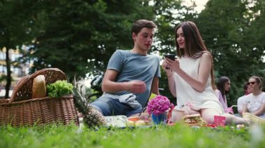 Gülümseyen mutlu genç çift parkta dinleniyor, piknik yapıyor, içki içiyor, kadın kızı seviyor, yaz kadını güzel, duygusal, açık havada eğleniyor, yavaş çekimde eğleniyor.