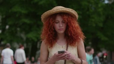 Yaklaş, kıvırcık saçlı, şapkalı çekici genç kadın akıllı telefon kullan, sokaklarda yürü, gülümse. Mutlu, yavaş çekimde yaz tatili portresi, günlük, beyaz, güzel kadın dış görünüşü.