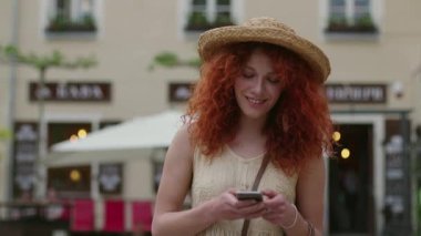 Kıvırcık saçlı, kırmızı saçlı, gülümseyen genç bir kadın şehir sokaklarında yürüyen akıllı bir telefon kullanıyor mutlu, yavaş çekimde yaz yüzü teknolojisi gelişigüzel, beyaz, güzel bir kadın.