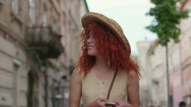 Şık, kıvırcık saçlı, şapkalı, şık bir genç kadın. Şehir sokaklarında yürürken akıllı telefon kullanıyor. Gülümsüyor, yaz aylarında gülümsüyor.