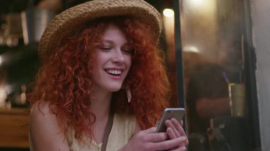 Yüzü gülümseyen, şapkalı, kızıl saçlı, güzel bir kadın kafede oturuyor. Moda kızı bağlantısını kullanıyor. Kafkasyalı akıllı telefonlu kadın.