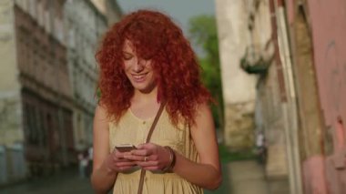 Kızıl saçlı genç bir kadın şehir sokaklarında yürüyor. Telefonla kameraya bak. Gülümse. Mutlu yaz gülümsemesi.