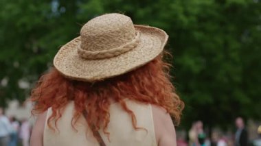 Çekici bir portre, şapkalı, kırmızı kıvırcık saçlı, gülümseyen, mutlu, yazın sokaklarda yürüyen beyaz tenli, güzel, açık hava manzaralı, sevimli, yavaş çekimde bir bayan.