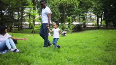 Güzel mutlu Afrikalı kadın piknik battaniyesinde oturuyor. Koca ve küçük kızı yaklaşıyor. Piknik. Açık havada. Gündüz vakti. Yaz.