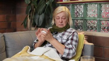 Mutsuz yaşlı kadın masaya kağıt havlu koyuyor. Gri kanepede battaniyenin altında yatan yaşlı kadın portresi. Yüksek ateş. Yakın plan. Kapalı.