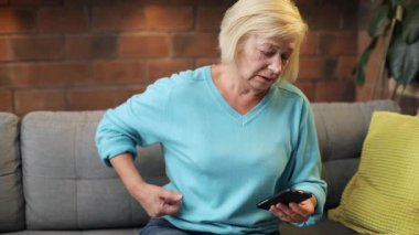 Sarı yastıkla gri koltukta oturan, akıllı telefon kullanan beyaz bir kadın. Mavi kazak ve kot pantolon giyen sevimli hasta büyükanne. Sırt ağrısı. - Eve. Oturma odası.