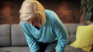 Bacağı ağrıyan beyaz bir kadın gri koltukta oturuyor, masaj yapıyor. Mavi kazak ve kot pantolon giyen sevimli yaşlı kadın portresi. İçeride. Oturma odası.
