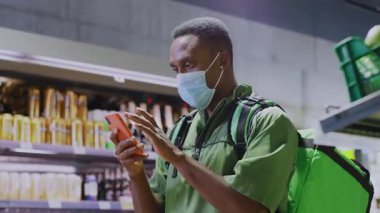Solunum maskesi takan Afro-Amerikan genç kurye telefon kullanarak müşteri arıyor. Süpermarket. Teslimat servisi. Karantina,